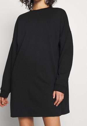 Kvinde iført en løs, langærmet sort sweaterkjole med rund halsudskæring og ribkanter mod en ensfarvet baggrund.