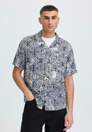 Junger Mann mit kurzen Haaren, trägt ein kurzärmeliges Hemd mit navyblauem und weißem Muster über einem weißen T-Shirt und schwarze Hose.