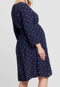 Mörkblå klänning med långa ärmar, blommönster, samlad midja och mjukt tyg. Kjolen slutar strax ovanför knäet och har en svag A-linjeform.