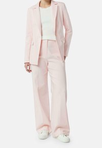 Completo su misura rosa chiaro con giacca aderente e pantaloni a gamba larga, abbinato a un top bianco a coste e sneaker bianche.