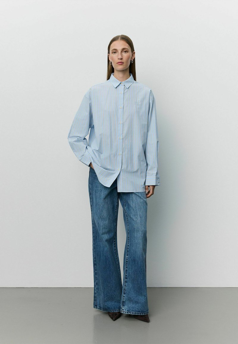 Femme aux longs cheveux raides porte une chemise oversize rayée bleu clair et un jean large bleu, debout contre un mur blanc uni.