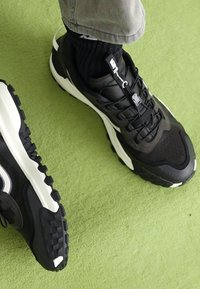 Black and white athletic shoes with a textured upper, lace-up design, and a thick, rugged sole, placed on a green surface.