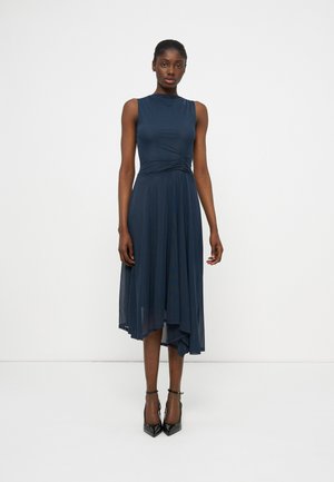 Γυναίκα που φοράει αμάνικο midi φόρεμα σε ναυτικό μπλε με ασύμμετρο τελείωμα και σούρα στη μέση, στέκεται μπροστά σε ένα απλό λευκό φόντο.