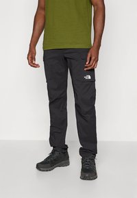 Μαύρο παντελόνι cargo με μπροστινές τσέπες, ελαφρύ ύφασμα, ελαστική μέση και μικρό λευκό λογότυπο στο μηρό· φοριέται με πράσινο πουκάμισο και ανθεκτικά παπούτσια.