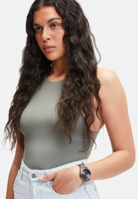 Olijfgroen mouwloos bodysuit gecombineerd met lichtblauwe hoge taille shorts. Het model draagt een zilveren horloge met een donkere wijzerplaat en een metalen band.