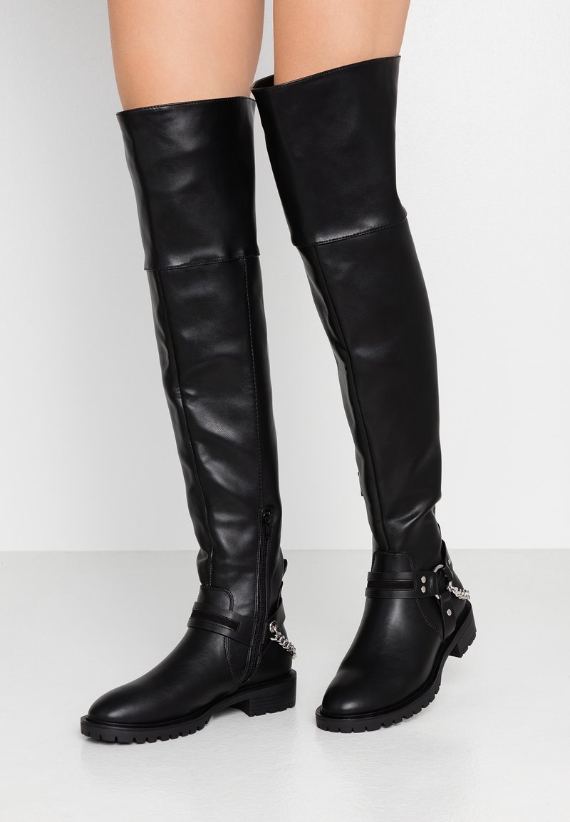 Stivali in pelle neri al ginocchio con suole chunky, cerniere laterali, fasce decorative e dettagli in catena argentata indossati sulle gambe.