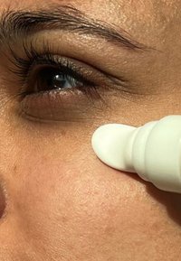 La punta bianca dell'applicatore per la cura della pelle tocca la pelle sotto un occhio marrone, mostrando una consistenza liscia con riflessi sottili e tonalità naturali.