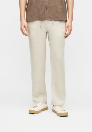 Pantalon beige à cordon de serrage porté avec des baskets blanches et une chemise marron à manches courtes boutonnée, visible de la taille aux pieds.