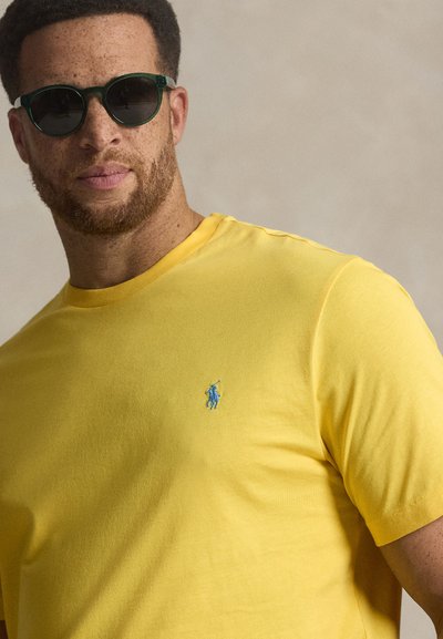 Κίτρινο βαμβακερό t-shirt με κοντά μανίκια, που έχει ένα μπλε κεντημένο λογότυπο στο στήθος. Συνοδεύεται από σκουρό πράσινα γυαλιά ηλίου.