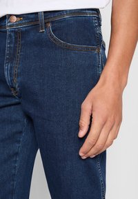 Donkerblauwe spijkerbroek van denim met contrasterende stiksel, een vijf-vak ontwerp en een metalen knoopsluiting bij de taille. Close-up view van de zijkant en hand.