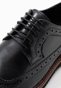 Chaussure brogue en cuir noir avec perforations wingtip, lacets noirs et semelle cousue marron, présentée en gros plan sur la partie supérieure avant.