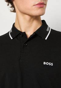 Jeune personne portant un polo noir avec des rayures blanches sur le col et le logo "BOSS" sur la poitrine.