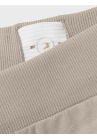 Cintura acanalada beige con una banda elástica blanca, que presenta un botón de plástico transparente y detalles de costura. Textura de tela suave.