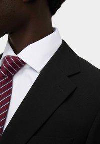 Zwarte colbert met een gladde textuur, witte overhemd en een bordeaux gestreepte das met blauwe accenten, zichtbaar etiket op het colbert.