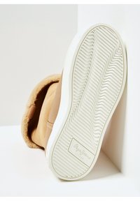 Brun ruskindsstøvle med foret i tan farvet imiteret pels, hvid tekstureret sål med bølget mønster og logo på siden.