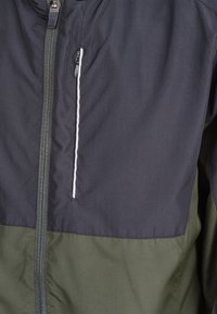 Svart och grön jacka med dragkedja fram och vita dekorativsömmar. Tillverkad av slät, lättviktig tyg. Har en bröstficka.