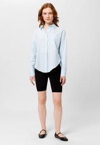 Camicia a righe blu chiaro e bianco con colletto arrotondato, abbinata a pantaloni corti neri da biker e ballerine nere. Tessuto liscio, fit contemporaneo.