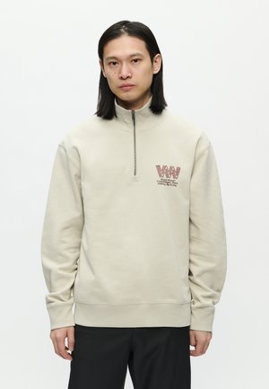 Junger Mann mit glattem schwarzen Haar trägt einen beigen Sweatshirt mit Viertel-Reißverschluss und rotem Logo sowie schwarze Hosen und steht vor einem schlichten weißen Hintergrund.