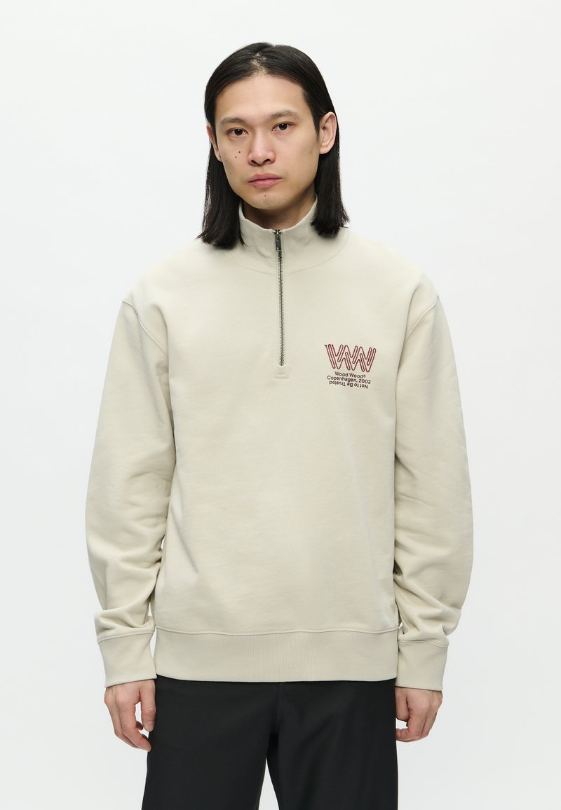 Giovane con capelli neri lisci, indossa una felpa beige a mezza zip con logo rosso e pantaloni neri, in piedi su uno sfondo bianco uniforme.