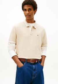 Beige Polohemd mit einem weißen Kragen, getragen über einem weißen Langarmhemd. Kombiniert mit blauen Jeans und einem braunen Gürtel mit Akzentfarben.