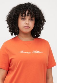 Narančasta pamučna T-shirt s kratkim rukavima, s bijelim izvezenim "Tommy Hilfiger" logom preko prsiju. Glatka tekstura, opušteni kroj.
