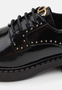 Chaussure en cuir noir poli avec des lacets noirs, des clous dorés le long du côté, une boucle dorée près du dessus, et une semelle épaisse texturée.