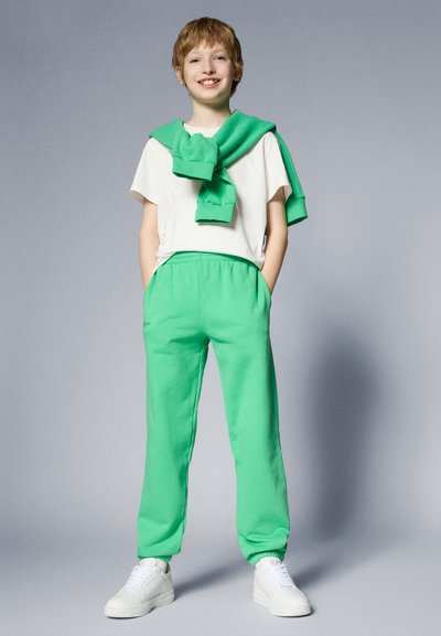 Pantalones de chándal verdes de algodón, con cintura elástica y bolsillos laterales, combinados con una camiseta blanca y un suéter verde caído sobre los hombros.