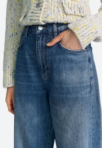 Svetlo modre džins hlače z visokim pasom in ravnim krojem, z sprednjim žepom in kontrastnim šivom. Kombinirane s teksturiranim kremnim pletenim puloverjem.