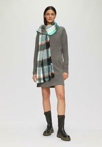 Robe grise à manches longues, écharpe à carreaux noir et blanc avec des touches d'orange et de teal, bottes noires à semelles épaisses, détails en tricot texturé.