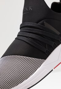 Lähikuva musta-valge tennisekinga detailist, millel on tekstuuriga varvas, mustad paelad ja valge tald punaste ja mustade aktsentidega.