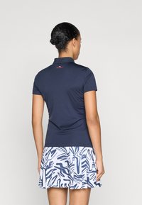 Jūras zilas polo krekls ar apkakli un īsām piedurknēm, apvienots ar plektu baltu svārku, kuram ir zils abstrakts raksts.