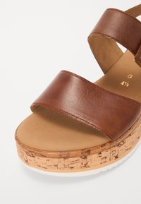 Brown leather sandal with two wide straps, cork platform with textured details, and a smooth tan insole. Outsole is white and flat.