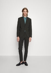 Dunkelgrüner Anzug mit slim fit, ausgestattet mit einem Zweiknopfjackett, Reverskragen und schmal zulaufenden Hosen. Kombiniert mit einem türkisfarbenen Hemd und schwarzen Slippern.