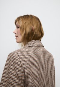 Blazer a motivo houndstooth marrone e crema con colletto a falda e spalle strutturate, caratterizzato da un tessuto testurizzato.