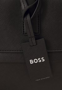 Sac en cuir noir avec une surface texturée, présentant une étiquette rectangulaire avec le mot "BOSS" en lettres blanches, attachée par une fine lanière.