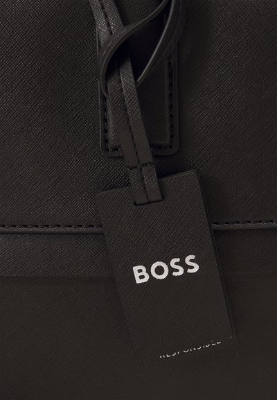 Sac en cuir noir avec une surface texturée, présentant une étiquette rectangulaire avec le mot "BOSS" en lettres blanches, attachée par une fine lanière.