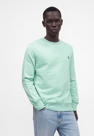 Homme portant un sweat-shirt vert clair à col rond avec un petit logo brodé sur la poitrine et un jean bleu clair, debout devant un fond blanc.