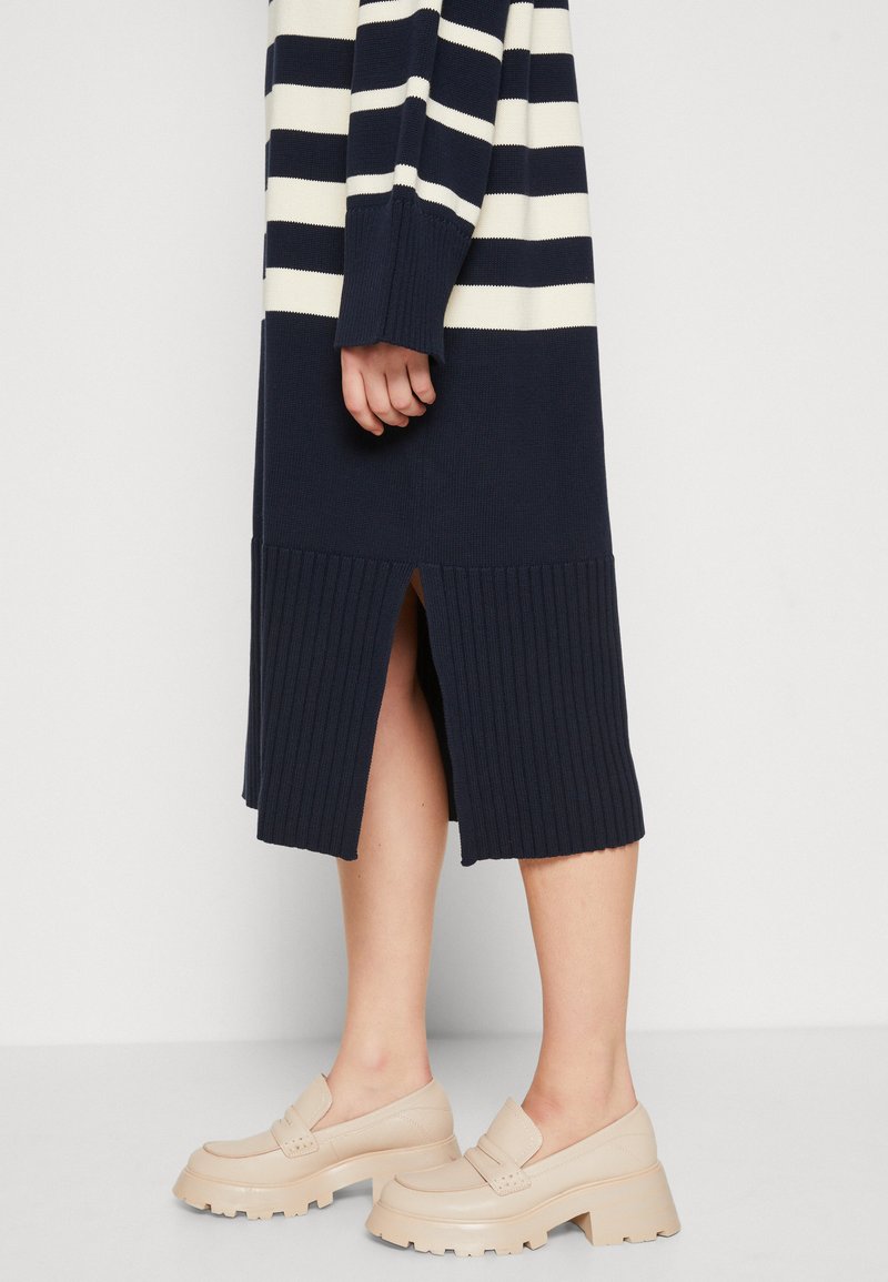 Granatowo-kremowa dzianinowa sukienka w paski z ribbowym wykończeniem i bocznym rozcięciem, zestawiona z jasnobeżowymi masywnymi mokasynami. Gładka textura na całej powierzchni.