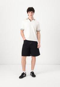 Ljust beige polo med bruna detaljer, i kombination med svarta shorts och svarta sneakers med vita ränder. Kort ärm, avslappnad passform.