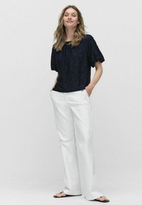 Marineblå blomsterMønstrede bluse med korte puffærmer, parret med hvid brede bukser og sorte sandaler, hænderne i lommerne.