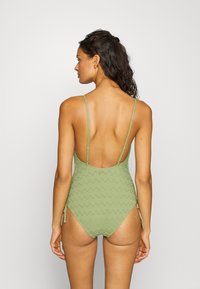Maillot de bain une pièce vert olive avec un motif en zigzag, bretelles fines réglables et un dos plongeant. Tissu doux et texturé.