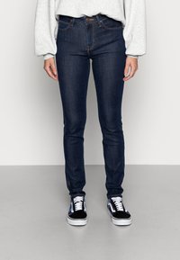 Vaqueros skinny de mezclilla en azul oscuro con costuras naranjas en contraste, con un diseño estándar de cinco bolsillos y cierre de botón. Combinados con zapatillas negras.