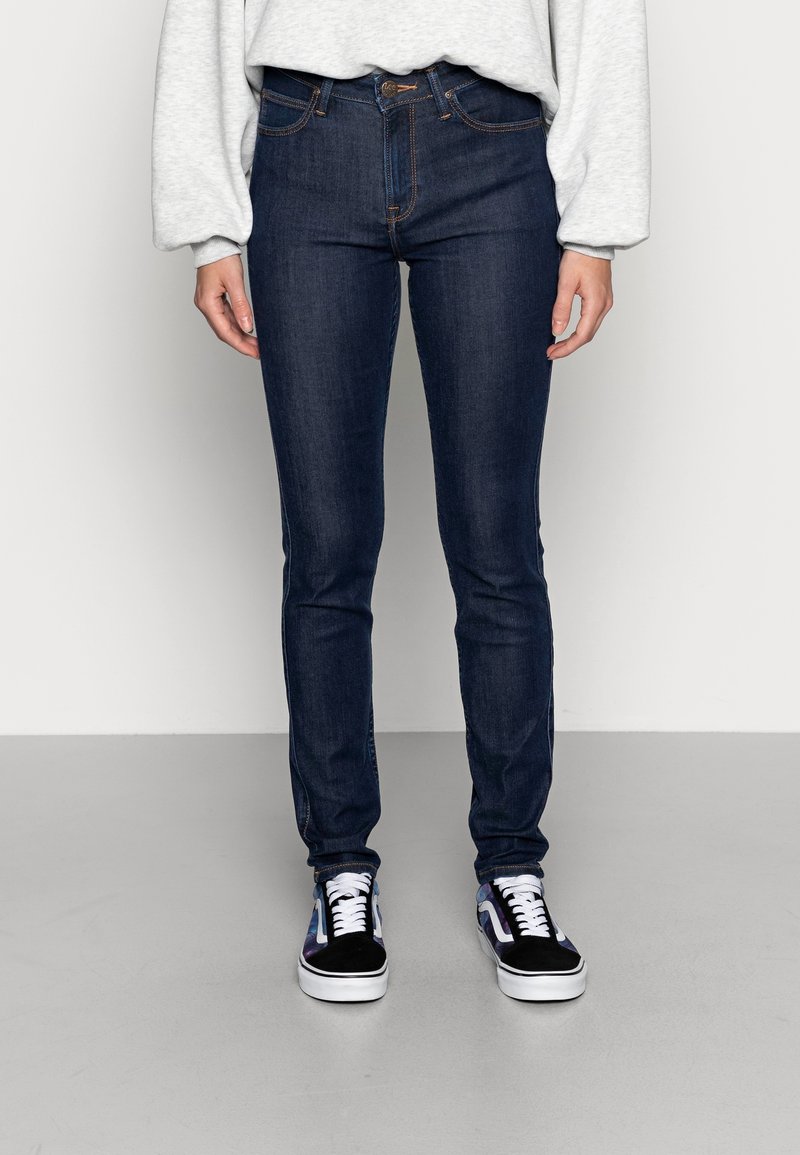 Vaqueros skinny de mezclilla en azul oscuro con costuras naranjas en contraste, con un diseño estándar de cinco bolsillos y cierre de botón. Combinados con zapatillas negras.