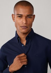 Camicia blu navy con chiusura a bottoni, design aderente, tessuto morbido e logo ricamato sobrio sul petto. Colletto strutturato e polsini con bottoni.