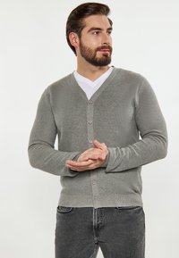 Szürke kötött kardigán V-nyakkal, gombos elülső résszel és bordás mandzsettákkal, fehér pólóval és sötét farmerral párosítva. Simaság jellemzi.