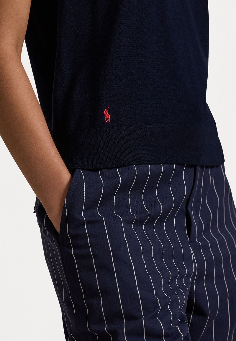 Granatowy sweter z krótkim rękawem, z małym czerwonym haftowanym logo. W połączeniu z granatowymi spodniami w paski z białymi liniami.