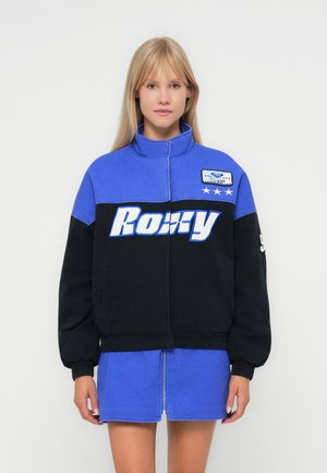 Blue and black jacket with a high collar, featuring a front zipper, large "Romy" text, and a patch on the chest. Soft fabric texture.