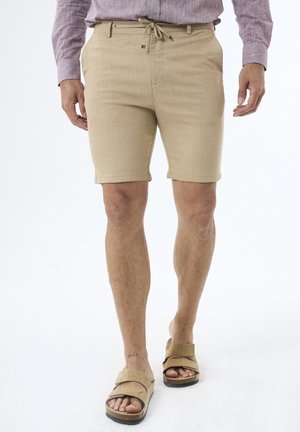 Shorts beiges en tissu texturé, longueur mi-cuisses, taille à cordon, et poches latérales, associés à des sandales à lanières marron.