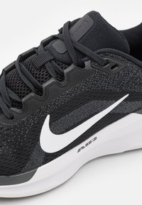 Chaussure de sport noire avec une tige en maille, un tissu texturé et un logo swoosh blanc bien visible. Elle dispose d'une semelle rembourrée et d'un design à lacets.