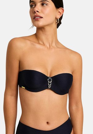 Femme portant un haut de bikini noir sans bretelles avec un détail décoratif en métal et tissu au centre, assorti à un bas de bikini coordonné.